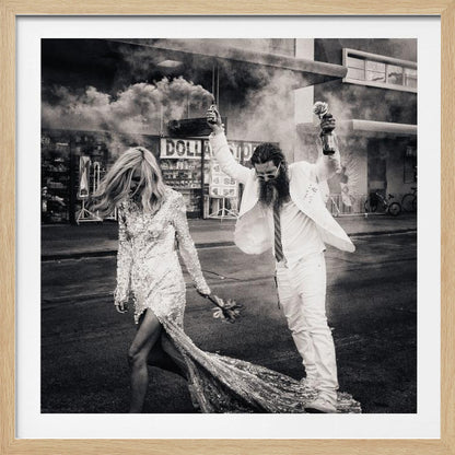 A black and white photograph in a light wood frame showing a stylish couple walking down a street. The woman wears a sparkling sequin gown, and the man, in a white suit with a long beard, triumphantly raises his arms, holding a smoke bomb and a bottle with a rose. Decor