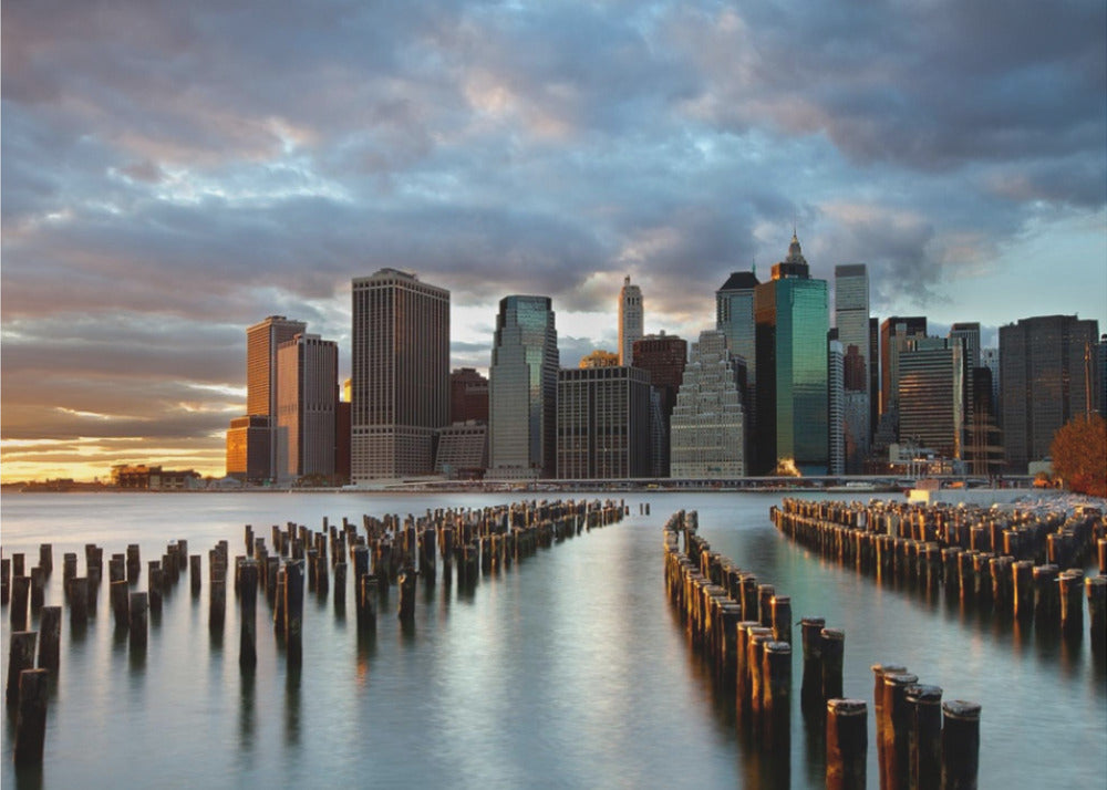 A framed photograph of the Lower Manhattan skyline at sunset, viewed from across the water. Rows of old wooden pier pilings lead the eye towards the city's skyscrapers, which are illuminated by the warm light of the setting sun under a dramatic, cloudy sky. Decor