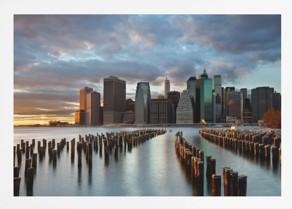 A framed photograph of the Lower Manhattan skyline at sunset, viewed from across the water. Rows of old wooden pier pilings lead the eye towards the city's skyscrapers, which are illuminated by the warm light of the setting sun under a dramatic, cloudy sky. Decor