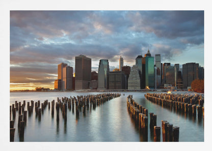 A framed photograph of the Lower Manhattan skyline at sunset, viewed from across the water. Rows of old wooden pier pilings lead the eye towards the city's skyscrapers, which are illuminated by the warm light of the setting sun under a dramatic, cloudy sky. Decor