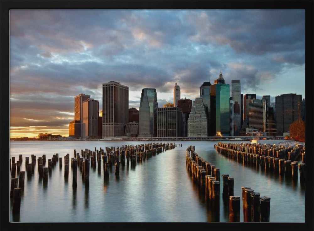 A framed photograph of the Lower Manhattan skyline at sunset, viewed from across the water. Rows of old wooden pier pilings lead the eye towards the city's skyscrapers, which are illuminated by the warm light of the setting sun under a dramatic, cloudy sky. Decor