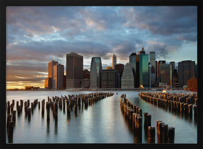 A framed photograph of the Lower Manhattan skyline at sunset, viewed from across the water. Rows of old wooden pier pilings lead the eye towards the city's skyscrapers, which are illuminated by the warm light of the setting sun under a dramatic, cloudy sky. Decor