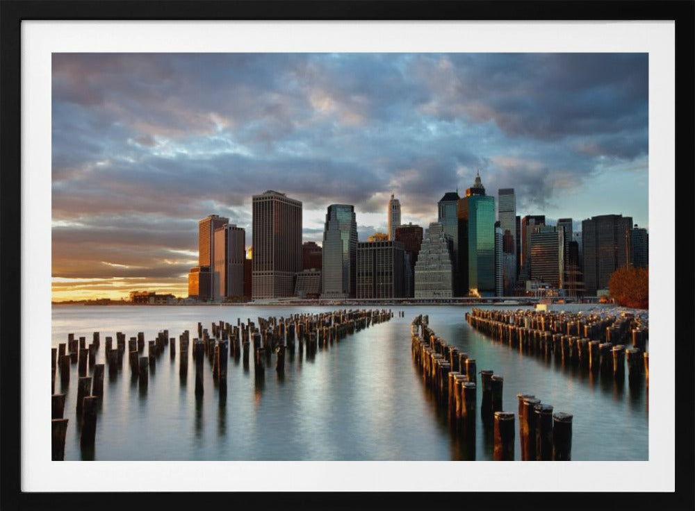 A framed photograph of the Lower Manhattan skyline at sunset, viewed from across the water. Rows of old wooden pier pilings lead the eye towards the city's skyscrapers, which are illuminated by the warm light of the setting sun under a dramatic, cloudy sky. Decor