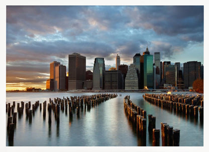 A framed photograph of the Lower Manhattan skyline at sunset, viewed from across the water. Rows of old wooden pier pilings lead the eye towards the city's skyscrapers, which are illuminated by the warm light of the setting sun under a dramatic, cloudy sky. Decor