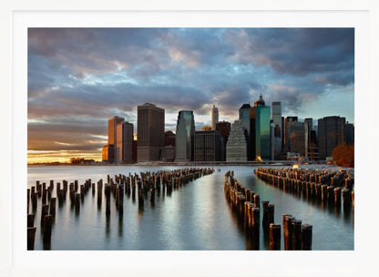 A framed photograph of the Lower Manhattan skyline at sunset, viewed from across the water. Rows of old wooden pier pilings lead the eye towards the city's skyscrapers, which are illuminated by the warm light of the setting sun under a dramatic, cloudy sky. Decor