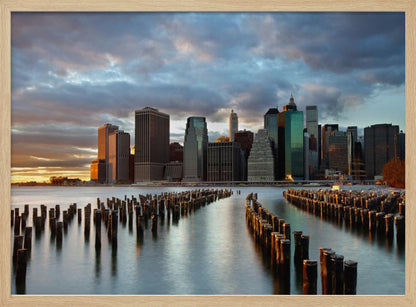 A framed photograph of the Lower Manhattan skyline at sunset, viewed from across the water. Rows of old wooden pier pilings lead the eye towards the city's skyscrapers, which are illuminated by the warm light of the setting sun under a dramatic, cloudy sky. Decor