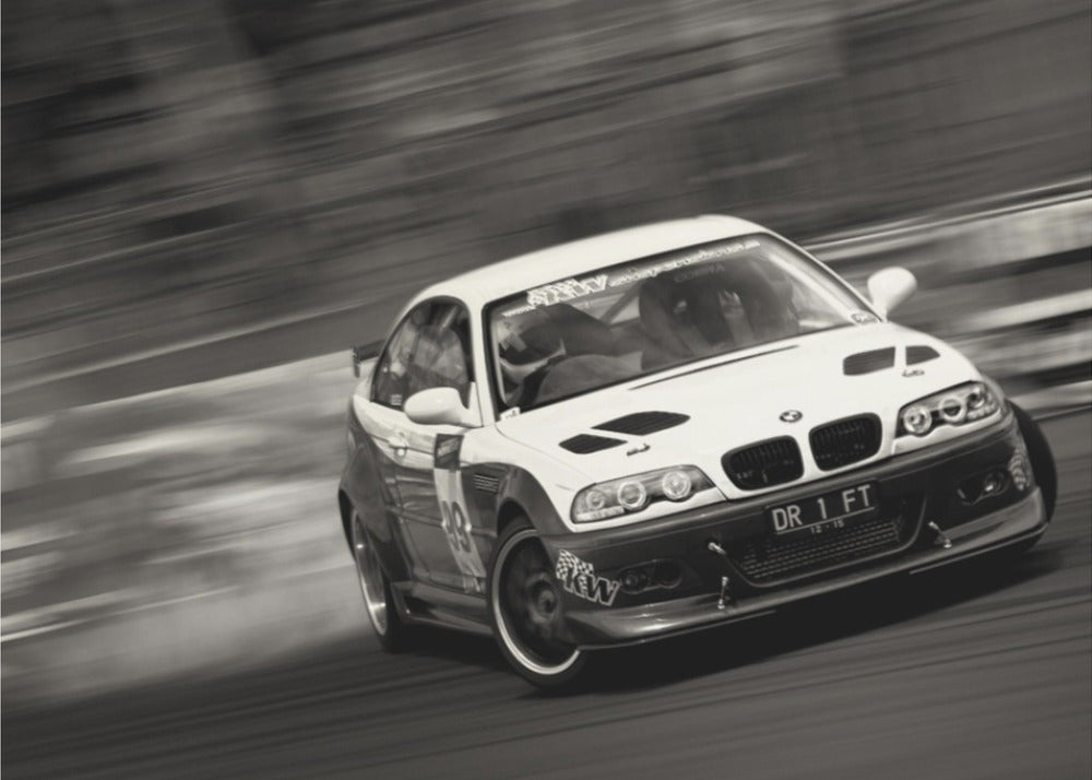 A framed, black and white action photograph of a white and black BMW race car drifting at high speed around a corner. The background is blurred with motion lines, emphasizing the car's speed as it turns, and a puff of smoke is visible from the tires. Wall Art