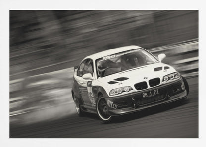 A framed, black and white action photograph of a white and black BMW race car drifting at high speed around a corner. The background is blurred with motion lines, emphasizing the car's speed as it turns, and a puff of smoke is visible from the tires. Wall Art