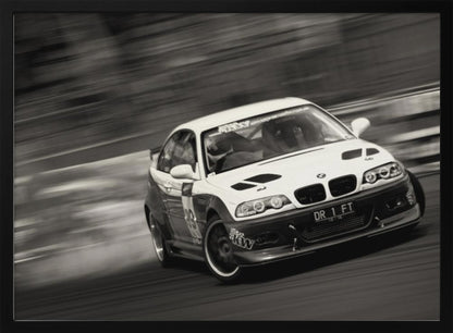 A framed, black and white action photograph of a white and black BMW race car drifting at high speed around a corner. The background is blurred with motion lines, emphasizing the car's speed as it turns, and a puff of smoke is visible from the tires. Wall Art