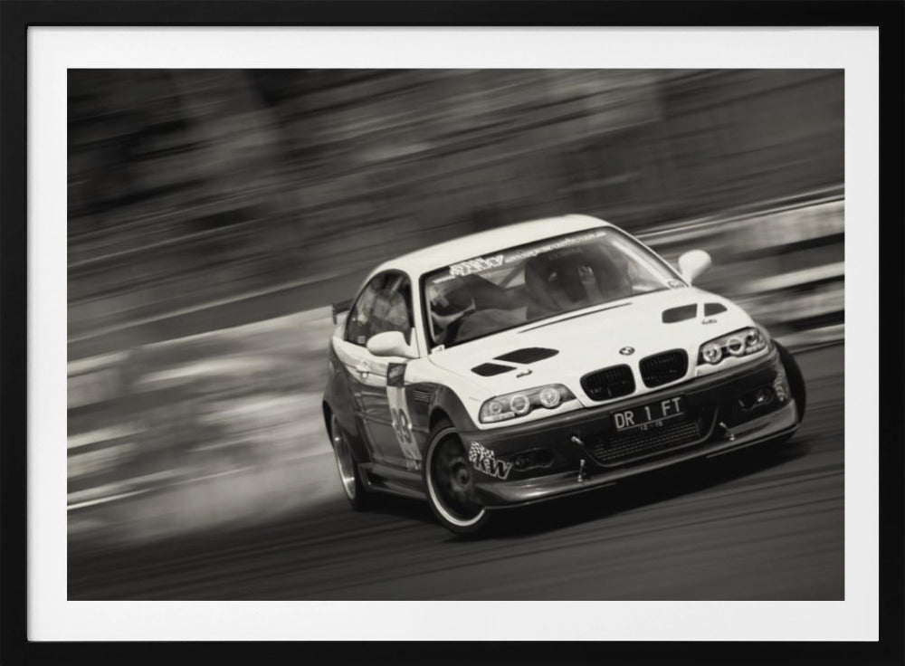 A framed, black and white action photograph of a white and black BMW race car drifting at high speed around a corner. The background is blurred with motion lines, emphasizing the car's speed as it turns, and a puff of smoke is visible from the tires. Wall Art