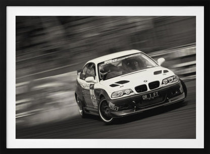 A framed, black and white action photograph of a white and black BMW race car drifting at high speed around a corner. The background is blurred with motion lines, emphasizing the car's speed as it turns, and a puff of smoke is visible from the tires. Wall Art