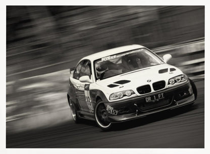 A framed, black and white action photograph of a white and black BMW race car drifting at high speed around a corner. The background is blurred with motion lines, emphasizing the car's speed as it turns, and a puff of smoke is visible from the tires. Wall Art