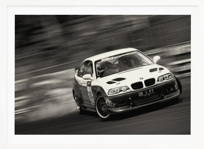 A framed, black and white action photograph of a white and black BMW race car drifting at high speed around a corner. The background is blurred with motion lines, emphasizing the car's speed as it turns, and a puff of smoke is visible from the tires. Wall Art