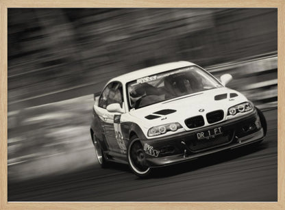 A framed, black and white action photograph of a white and black BMW race car drifting at high speed around a corner. The background is blurred with motion lines, emphasizing the car's speed as it turns, and a puff of smoke is visible from the tires. Wall Art