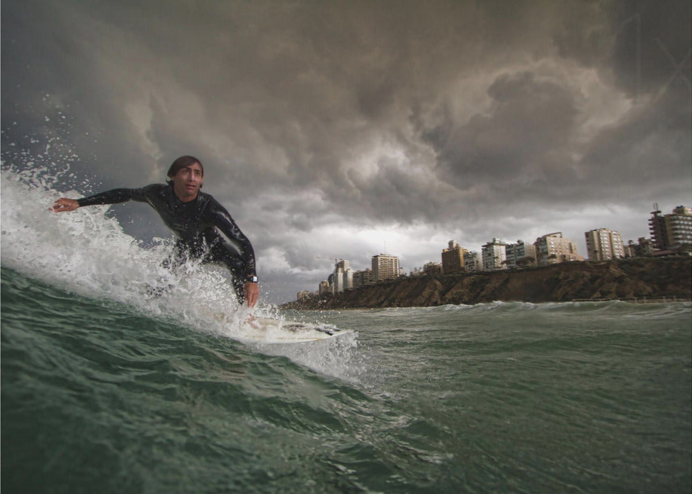 A close-up, low-angle shot of a male surfer in a black wetsuit riding a dark green ocean wave. In the background, a city skyline sits atop a cliff under a dramatic, dark, and stormy sky. The image is presented within a silver picture frame. Print