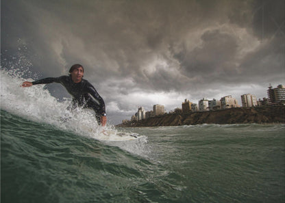 A close-up, low-angle shot of a male surfer in a black wetsuit riding a dark green ocean wave. In the background, a city skyline sits atop a cliff under a dramatic, dark, and stormy sky. The image is presented within a silver picture frame. Print