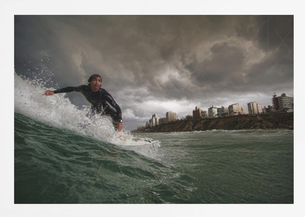 A close-up, low-angle shot of a male surfer in a black wetsuit riding a dark green ocean wave. In the background, a city skyline sits atop a cliff under a dramatic, dark, and stormy sky. The image is presented within a silver picture frame. Print