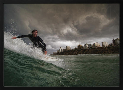 A close-up, low-angle shot of a male surfer in a black wetsuit riding a dark green ocean wave. In the background, a city skyline sits atop a cliff under a dramatic, dark, and stormy sky. The image is presented within a silver picture frame. Print