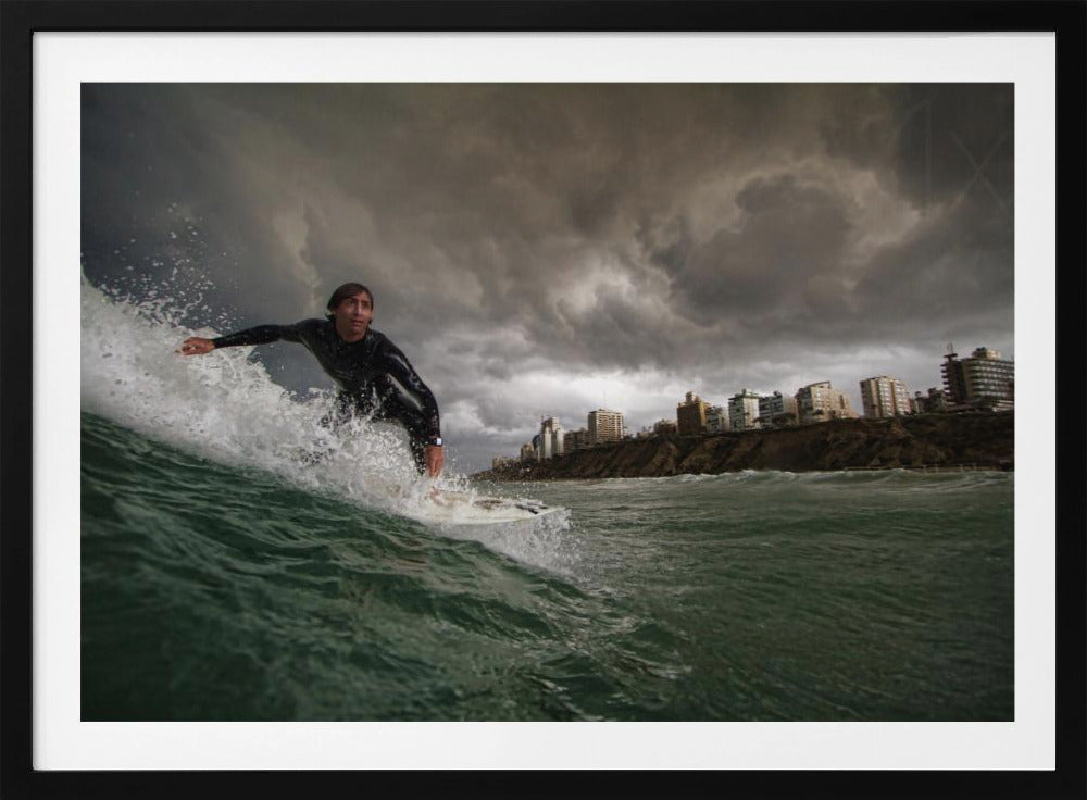 A close-up, low-angle shot of a male surfer in a black wetsuit riding a dark green ocean wave. In the background, a city skyline sits atop a cliff under a dramatic, dark, and stormy sky. The image is presented within a silver picture frame. Print