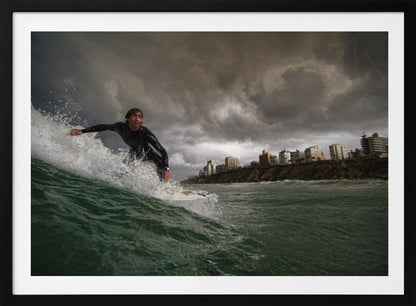 A close-up, low-angle shot of a male surfer in a black wetsuit riding a dark green ocean wave. In the background, a city skyline sits atop a cliff under a dramatic, dark, and stormy sky. The image is presented within a silver picture frame. Print