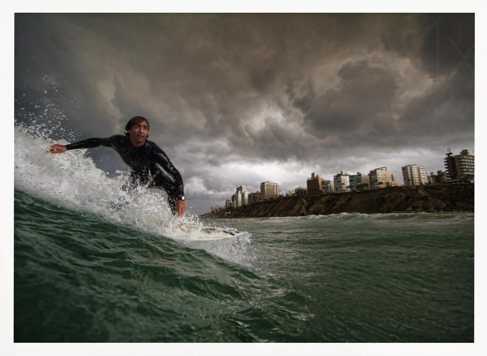 A close-up, low-angle shot of a male surfer in a black wetsuit riding a dark green ocean wave. In the background, a city skyline sits atop a cliff under a dramatic, dark, and stormy sky. The image is presented within a silver picture frame. Print