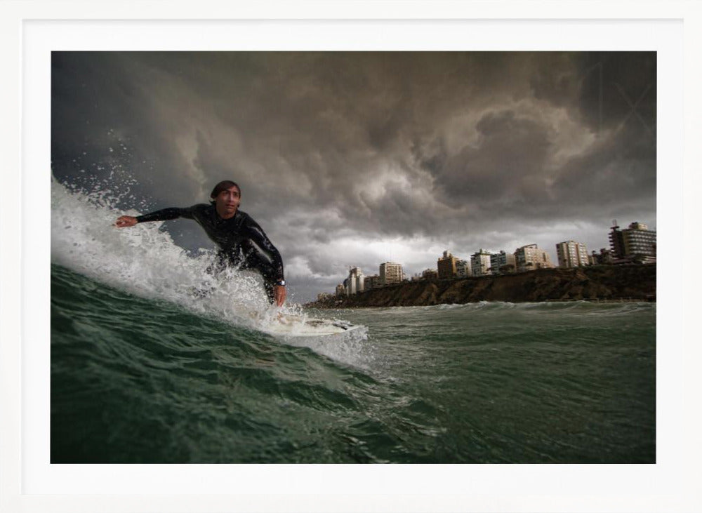 A close-up, low-angle shot of a male surfer in a black wetsuit riding a dark green ocean wave. In the background, a city skyline sits atop a cliff under a dramatic, dark, and stormy sky. The image is presented within a silver picture frame. Print
