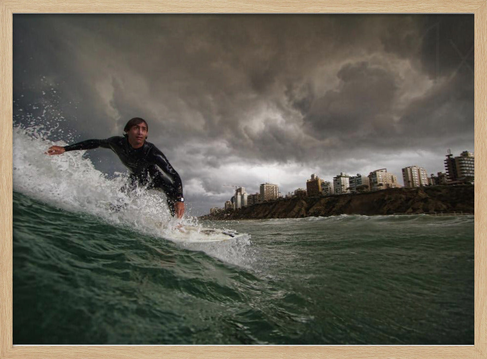 A close-up, low-angle shot of a male surfer in a black wetsuit riding a dark green ocean wave. In the background, a city skyline sits atop a cliff under a dramatic, dark, and stormy sky. The image is presented within a silver picture frame. Print