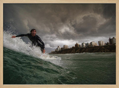 A close-up, low-angle shot of a male surfer in a black wetsuit riding a dark green ocean wave. In the background, a city skyline sits atop a cliff under a dramatic, dark, and stormy sky. The image is presented within a silver picture frame. Print