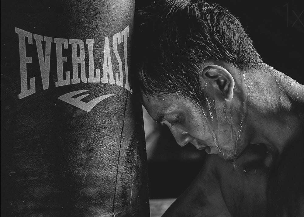 A dramatic black and white close-up photograph of an exhausted boxer resting his head against a heavy Everlast punching bag, with sweat dripping down his face and neck. Poster