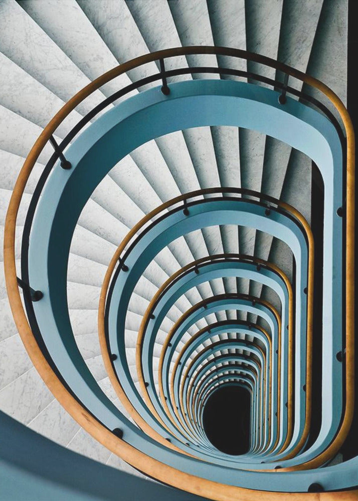 A high-angle, top-down photograph of a multi-story rectangular spiral staircase, creating a hypnotic, receding pattern. The steps are light gray marble, contrasted by shadows, and the balustrade is a striking light blue with a warm wooden handrail, leading down to a dark abyss at the center. Wall Art