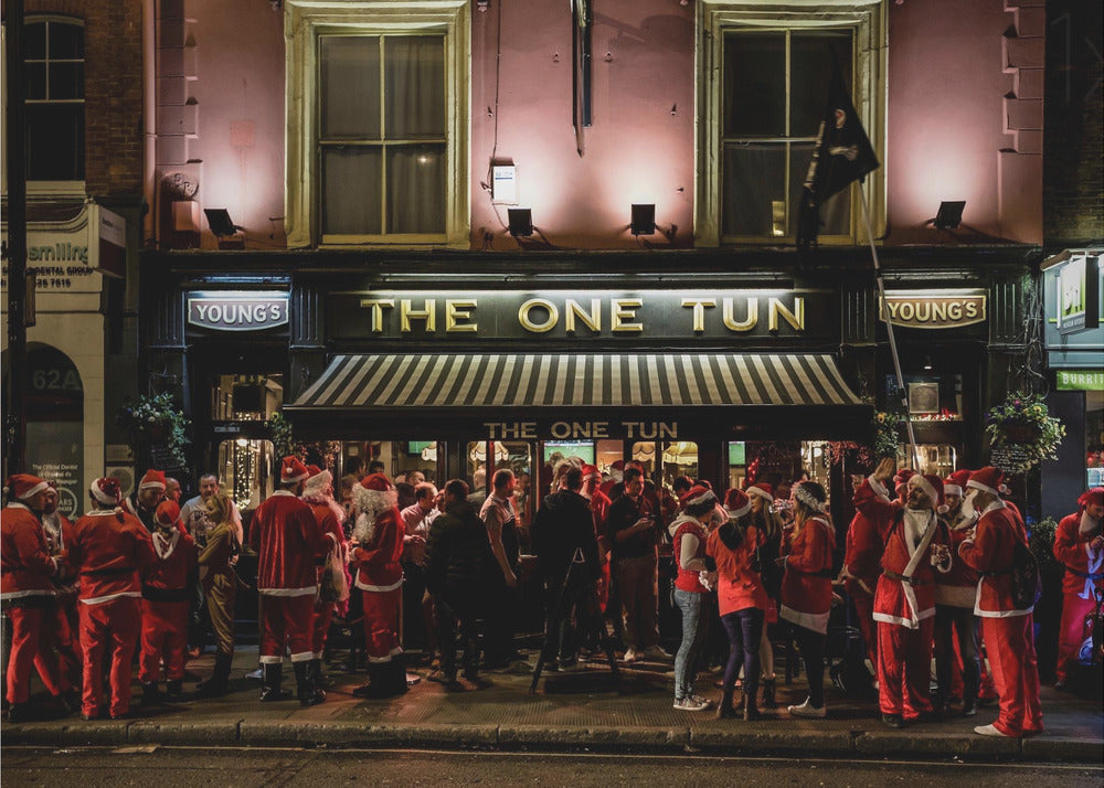 A nighttime photograph of a large crowd of people dressed in Santa Claus costumes standing outside a warmly lit pub called 'The One Tun', creating a festive holiday atmosphere on a city street. Print