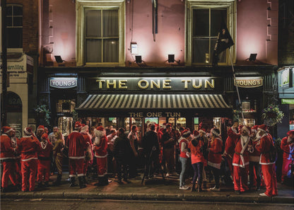 A nighttime photograph of a large crowd of people dressed in Santa Claus costumes standing outside a warmly lit pub called 'The One Tun', creating a festive holiday atmosphere on a city street. Print