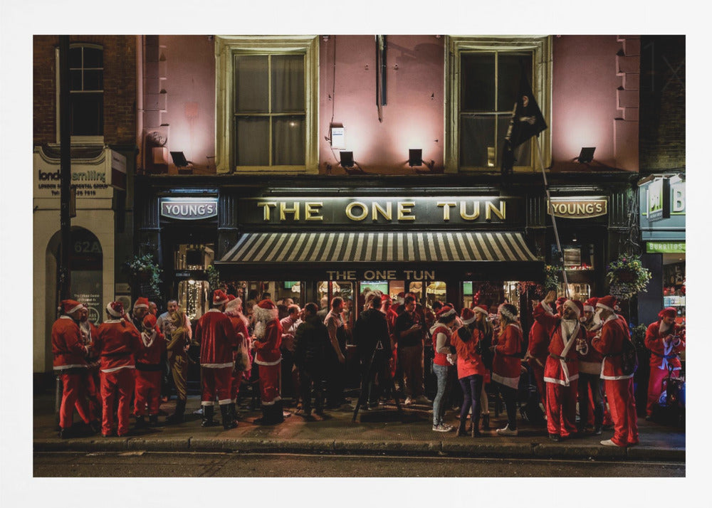 A nighttime photograph of a large crowd of people dressed in Santa Claus costumes standing outside a warmly lit pub called 'The One Tun', creating a festive holiday atmosphere on a city street. Print