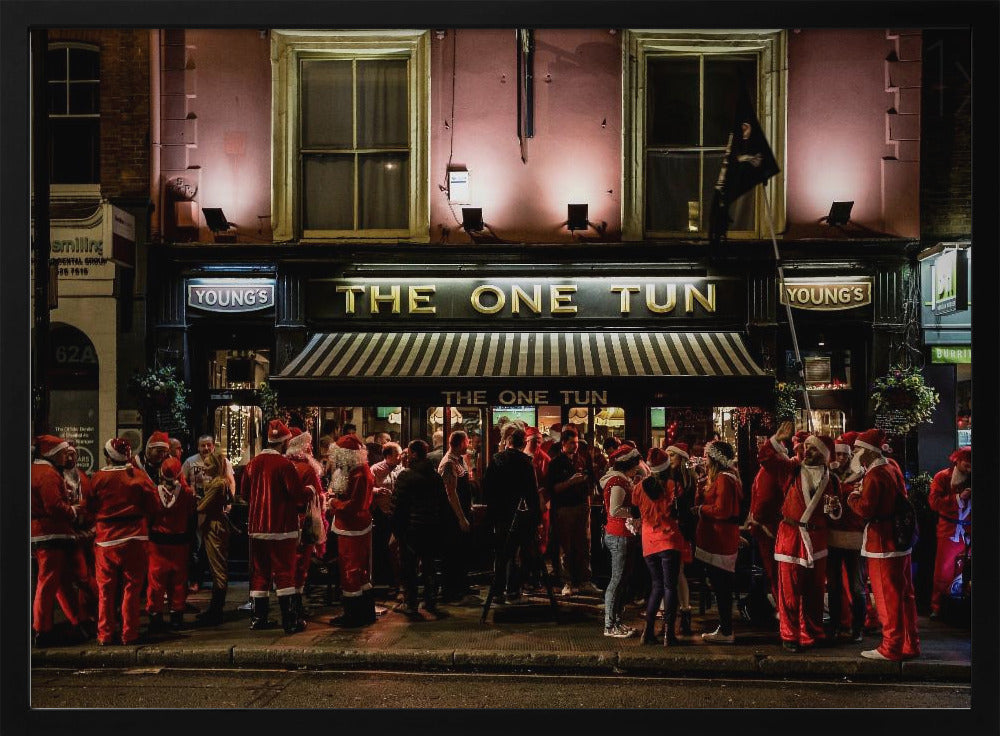 A nighttime photograph of a large crowd of people dressed in Santa Claus costumes standing outside a warmly lit pub called 'The One Tun', creating a festive holiday atmosphere on a city street. Print