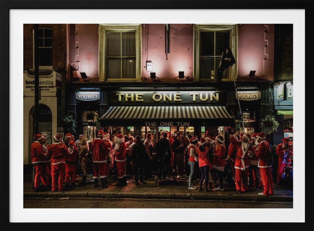 A nighttime photograph of a large crowd of people dressed in Santa Claus costumes standing outside a warmly lit pub called 'The One Tun', creating a festive holiday atmosphere on a city street. Print