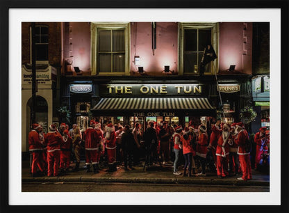 A nighttime photograph of a large crowd of people dressed in Santa Claus costumes standing outside a warmly lit pub called 'The One Tun', creating a festive holiday atmosphere on a city street. Print