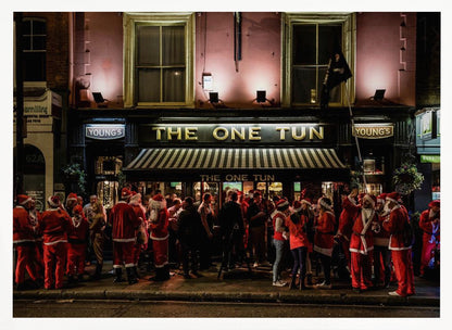 A nighttime photograph of a large crowd of people dressed in Santa Claus costumes standing outside a warmly lit pub called 'The One Tun', creating a festive holiday atmosphere on a city street. Print