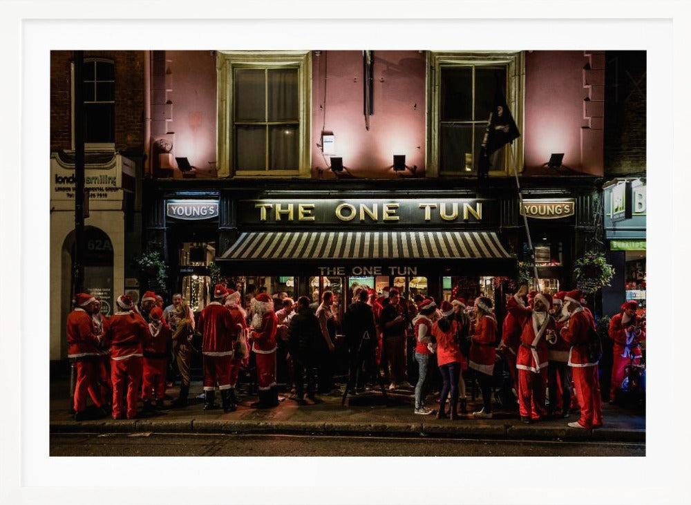 A nighttime photograph of a large crowd of people dressed in Santa Claus costumes standing outside a warmly lit pub called 'The One Tun', creating a festive holiday atmosphere on a city street. Print