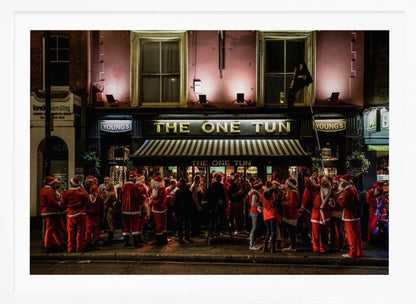 A nighttime photograph of a large crowd of people dressed in Santa Claus costumes standing outside a warmly lit pub called 'The One Tun', creating a festive holiday atmosphere on a city street. Print