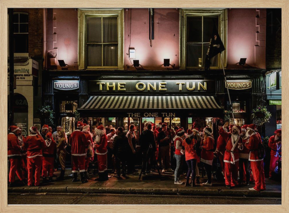 A nighttime photograph of a large crowd of people dressed in Santa Claus costumes standing outside a warmly lit pub called 'The One Tun', creating a festive holiday atmosphere on a city street. Print