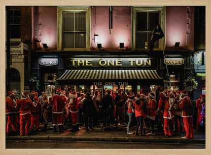 A nighttime photograph of a large crowd of people dressed in Santa Claus costumes standing outside a warmly lit pub called 'The One Tun', creating a festive holiday atmosphere on a city street. Print