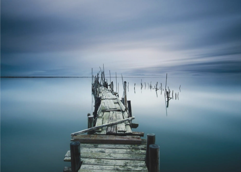 A framed, long exposure photograph of a weathered wooden pier extending into calm, misty blue water under a dramatic, cloudy sky. Wall Art