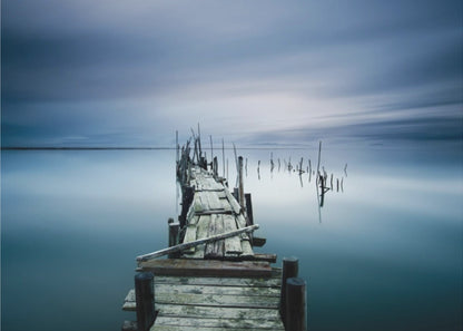 A framed, long exposure photograph of a weathered wooden pier extending into calm, misty blue water under a dramatic, cloudy sky. Wall Art