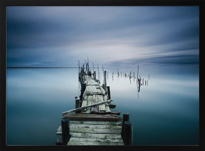 A framed, long exposure photograph of a weathered wooden pier extending into calm, misty blue water under a dramatic, cloudy sky. Wall Art
