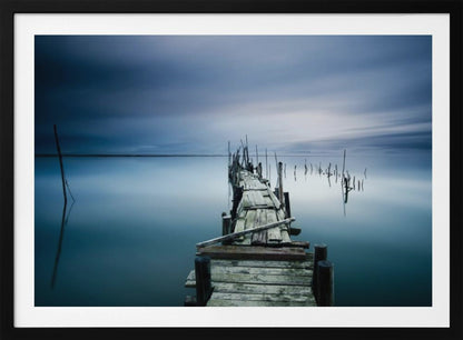 A framed, long exposure photograph of a weathered wooden pier extending into calm, misty blue water under a dramatic, cloudy sky. Wall Art