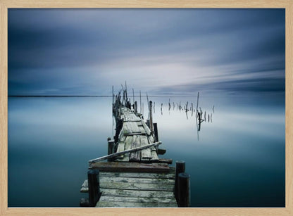 A framed, long exposure photograph of a weathered wooden pier extending into calm, misty blue water under a dramatic, cloudy sky. Wall Art