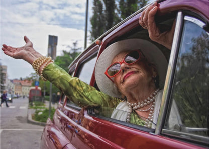 A stylish elderly woman wearing a white hat, red sunglasses, and pearl necklaces leans out of the passenger window of a classic red car. Her arm is outstretched with a joyful expression on her face, enjoying a ride through a city street. Poster