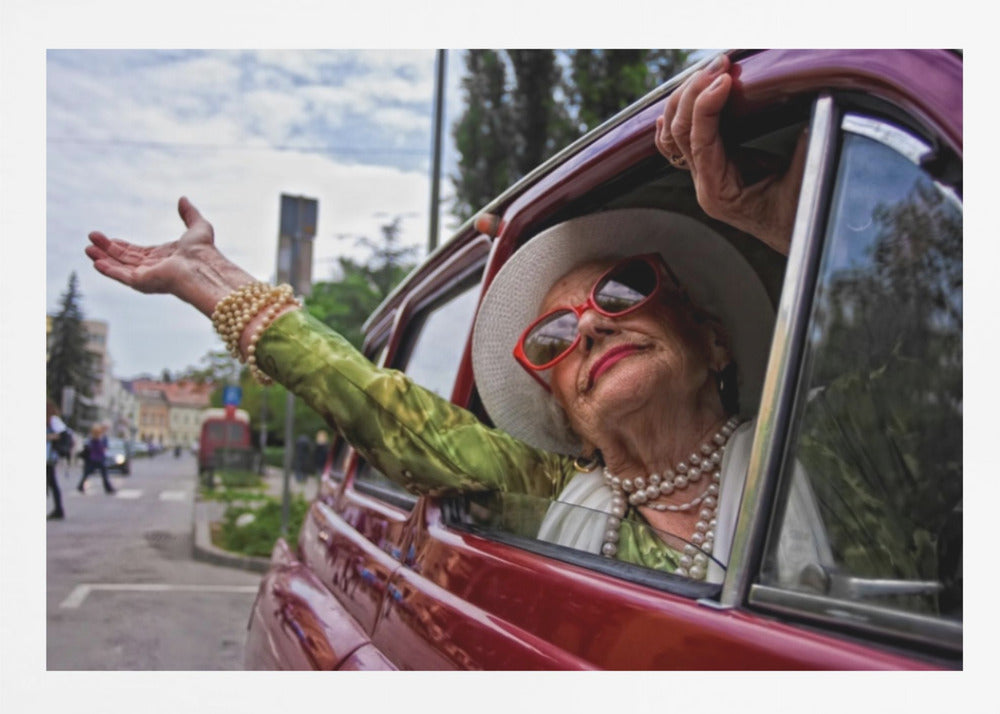 A stylish elderly woman wearing a white hat, red sunglasses, and pearl necklaces leans out of the passenger window of a classic red car. Her arm is outstretched with a joyful expression on her face, enjoying a ride through a city street. Poster