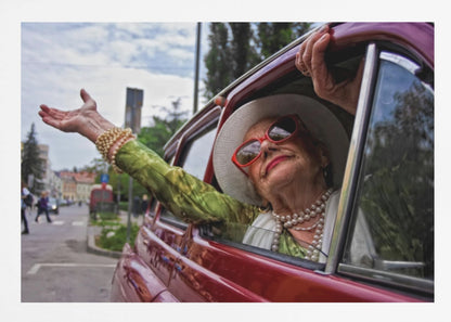 A stylish elderly woman wearing a white hat, red sunglasses, and pearl necklaces leans out of the passenger window of a classic red car. Her arm is outstretched with a joyful expression on her face, enjoying a ride through a city street. Poster
