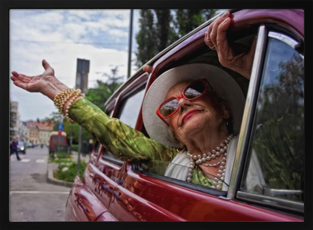 A stylish elderly woman wearing a white hat, red sunglasses, and pearl necklaces leans out of the passenger window of a classic red car. Her arm is outstretched with a joyful expression on her face, enjoying a ride through a city street. Poster